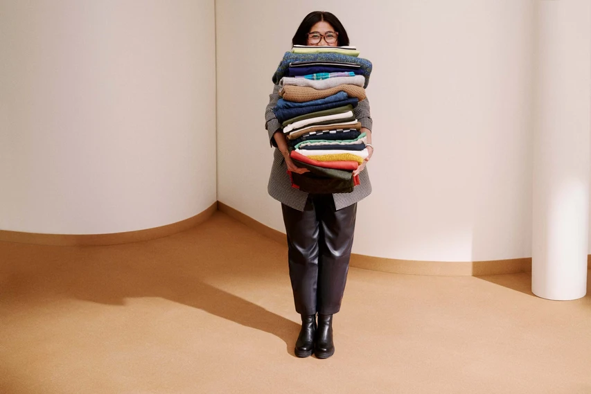 A professional photograph of Rowena holding a stack of clothing items from John Lewis. Peering over the top of the clothes and smiling.