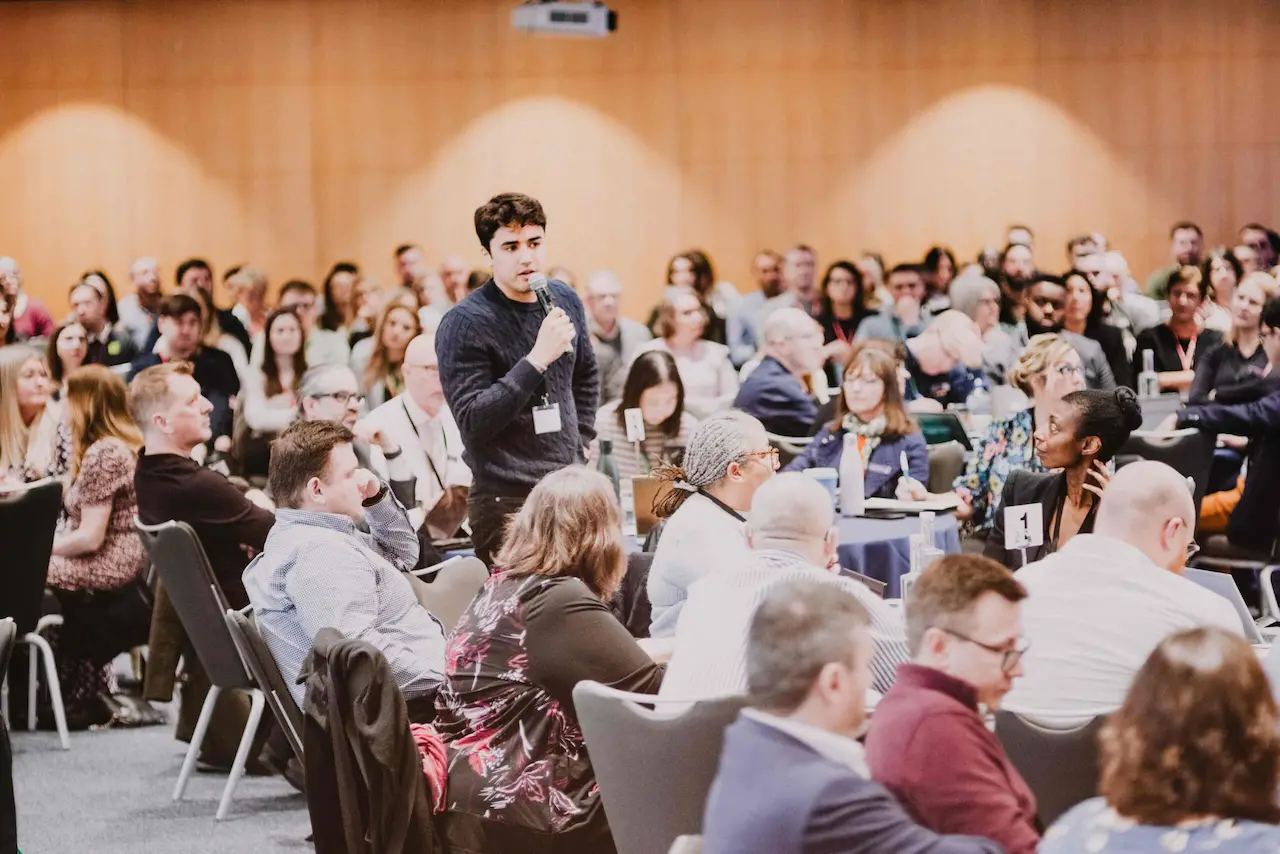 Large group of people sat around tables in big meeting