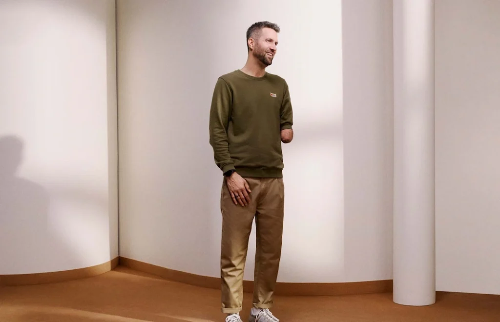 Tom at a professional photoshoot staring and smiling to the side wearing a green jumper and beige trousers. 